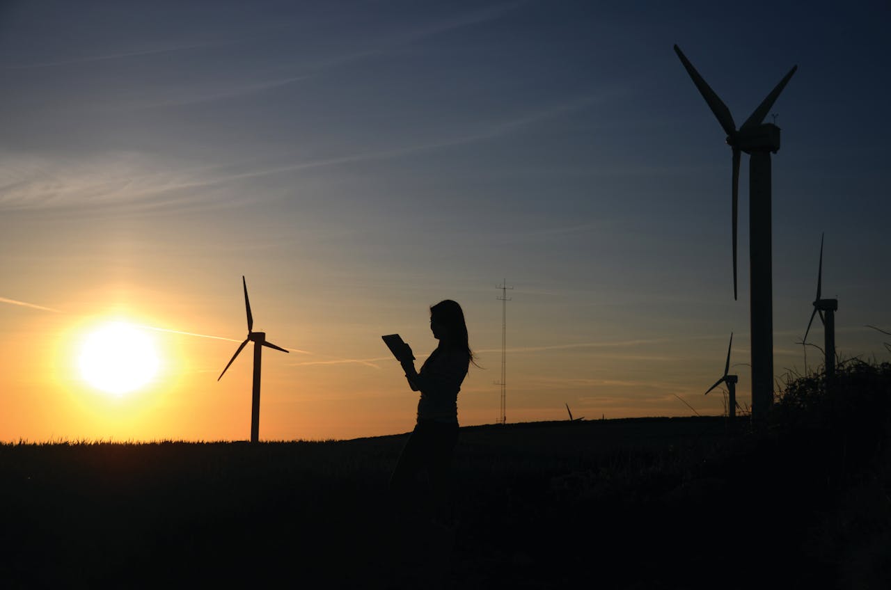 A silhouette of a woman using a device near wind turbines at sunset, representing renewable energy.